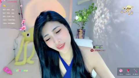 Media: A video of an Asian woman with long black hair, wearing a blue halter top, smiling in a bright room with a potted plant and a yellow figurine in the background.