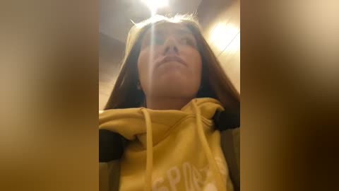 Media: Video of a young girl with light brown hair, wearing a yellow hoodie, looking up. The background is blurred, suggesting an indoor setting with warm lighting.