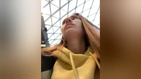 Media: A video of a young woman with long, straight blonde hair, wearing a yellow hoodie, standing indoors under a glass dome ceiling. The image is slightly blurred, with a focus on her head and shoulders.