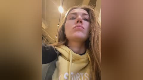 Media: Video of a young woman with long brown hair, wearing a yellow hooded sweatshirt with black sleeves, indoors. She has a neutral expression. Background shows a dimly lit room with exposed ceiling pipes and a hanging light fixture.