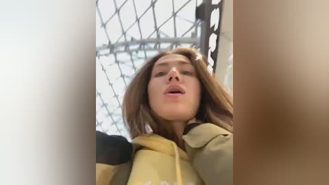 Media: A video of a young woman with long brown hair, fair skin, and parted lips, wearing a beige coat. She is standing against a wire fence in a snowy, industrial background.