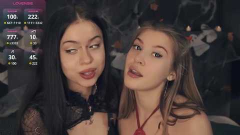 Media: Video of two young women with fair skin, one with long black hair, the other with straight blonde hair. They wear red lipstick and black lace tops. Background shows a dark, dimly-lit room with a TV screen displaying stats.