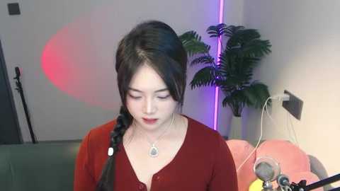 Media: Video of an Asian woman with fair skin, long black hair, and a red top, seated in a studio with a potted plant, pink lighting, and a microphone.