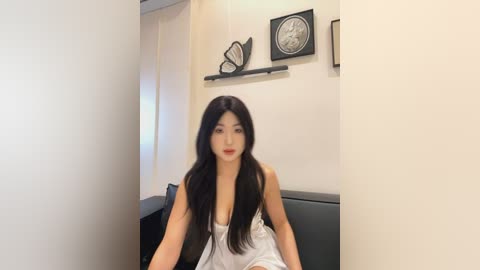 Media: Video of an Asian woman with long black hair, wearing a white lace dress, sitting on a black chair in a minimalist room with white walls, black shelves, and butterfly decor.