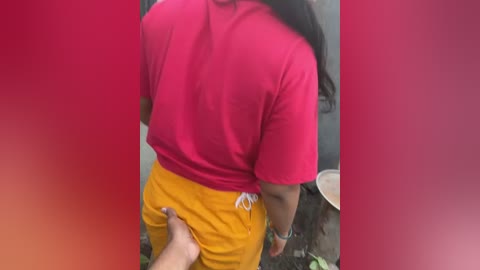 Media: Video of a person wearing a red shirt and mustard-yellow shorts, with a hand grabbing their buttocks. Background is blurred, featuring a red gradient overlay.