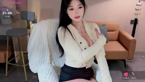 Media: Video of an East Asian woman with long black hair, wearing a low-cut cream blouse and black skirt, seated on a beige couch, in a modern living room.