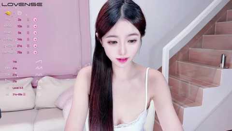 Media: Video of an Asian woman with long, straight black hair, fair skin, and pink lipstick, wearing a white lace camisole, sitting on a beige sofa in a pastel-pink room.