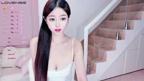 Media: Video of an East Asian woman with long black hair, fair skin, and pink lipstick, wearing a white tank top, sitting indoors near a white staircase.