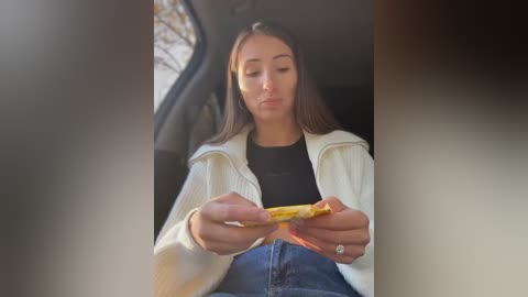 Media: Video of a young woman with long brown hair, wearing a white jacket and black top, eating a slice of pizza while seated in a car, window view of blurred trees.