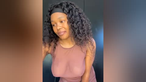 Media: Video of a smiling African-American woman with a medium-dark skin tone and curly black hair, wearing a sheer mauve top revealing her breasts, against a blurred, dark background.