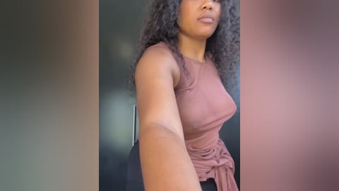 Media: Video of a Black woman with curly hair in a sleeveless mauve top, standing indoors against a dark background, with a blurred, neutral-toned gradient behind her.