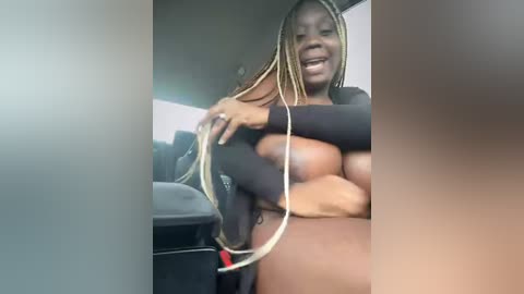 Media: A video of a smiling Black woman with long, blonde braids, wearing a black top, partially covering her breasts, sitting in a car. The background shows a blurred indoor setting.