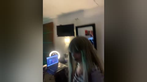 Media: A blurry video shows a young woman with long, dyed hair, standing in a dimly lit room with a laptop on a table.