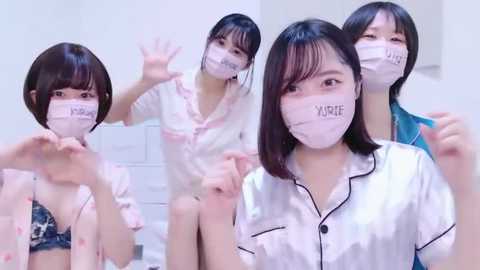 Media: Video of four young East Asian women in white satin robes and masks, posing in a white-tiled bathroom. They smile and wave, with one holding a phone.