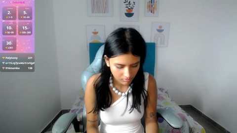 Media: Video of a young Latina woman with straight black hair, wearing a white sleeveless top and a pearl necklace, sitting on a light blue gaming chair in a minimalistic bedroom with white walls and abstract art.