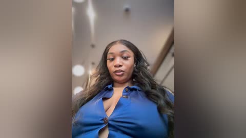 Media: Video of a plus-size Black woman with long, wavy hair, wearing a blue button-up shirt. She has a full-figured body and a confident expression. The background is blurred, suggesting an indoor setting with soft lighting.