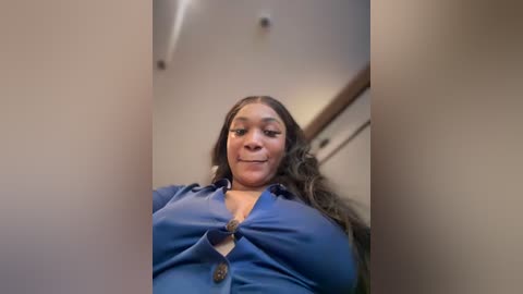 Media: A video of a Black woman with long, wavy hair, wearing a blue button-up blouse, standing in a dimly lit room with beige walls and recessed lighting.