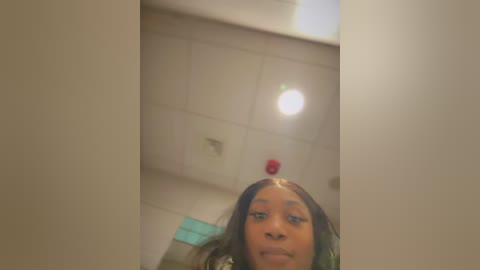 Media: Video of a young Black woman with medium brown skin, wearing a red beanie, taken indoors with a blurred, beige background featuring a ceiling light and white tiles.