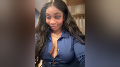 Media: A video of a smiling, dark-skinned woman with long, wavy black hair in a blue, open-collared blouse. The background features a blurred staircase and beige walls.