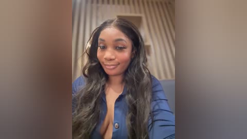 Media: Video of a Black woman with long, wavy, dark hair, wearing a blue button-up shirt, smiling, standing in a beige hallway with vertical stripes.