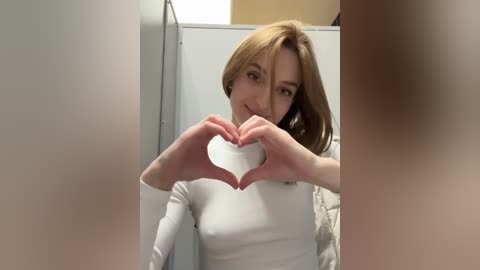 Media: A video of a young woman with light skin and shoulder-length blonde hair, smiling and forming a heart shape with her hands in a bathroom. She wears a white top.