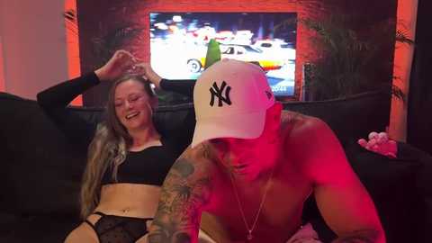 Media: Video of a smiling woman with long hair in a black crop top and high-waisted pants sitting next to a shirtless man with a white New York Yankees cap, tattoos, and a red glow.