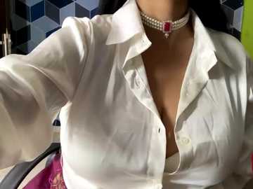 Media: A video of a woman's upper torso, wearing a white button-up shirt, revealing cleavage, adorned with a pearl choker and pink blazer. Background features a geometric patterned wall.