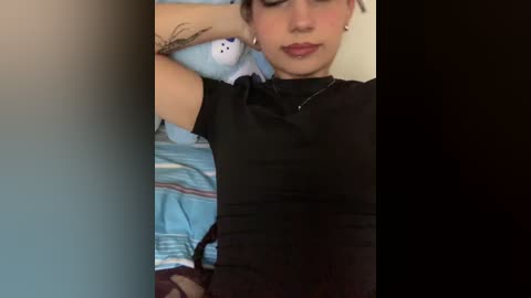 Media: A video of a young woman with light skin, medium-length brown hair, and wearing a black top, lying on a bed with a blue and white striped blanket. She has a tattoo on her right forearm and is resting her head on a stuffed animal.