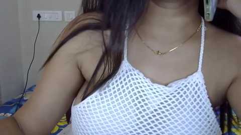 Media: Video of a young woman with long brown hair, wearing a white crochet halter top, gold necklace, and a colorful bedspread in the background.