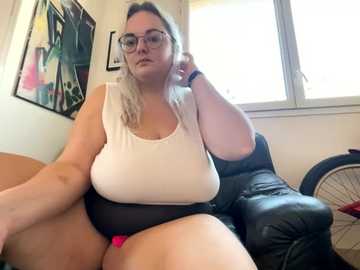 Media: Video of a plus-sized, light-skinned woman with glasses and blonde hair, wearing a white tank top and black shorts, seated on a black leather couch. Background features a window, colorful artwork, and a fan.