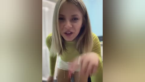 Media: Video of a young blonde woman with light skin, wearing a green long-sleeve top and white shorts, leaning forward, her tongue sticking out, blurry background.