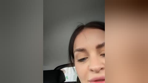 Media: Video of a young woman with dark hair and fair skin, partially visible, leaning on a black car seat, with a beige wall in the background.