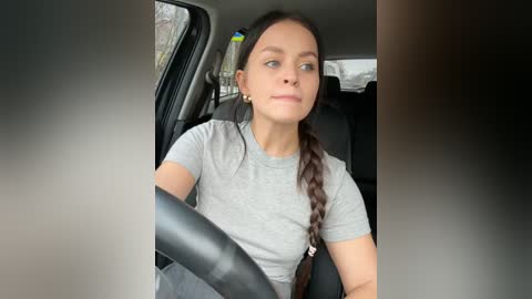 Media: Video of a young woman with light skin, brown hair in a braid, wearing a gray t-shirt, driving a car with a black steering wheel and seat, background shows a blurred exterior.