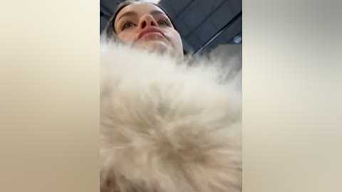 Media: Video of a woman from below, showing her face and upper body. She has fair skin, brown hair, and is wearing a white fur coat. The background is blurred, suggesting an indoor setting with a grey ceiling.