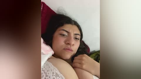 Media: Video of a young woman with medium skin tone, dark hair, and a curvy physique lying on a bed, partially covered by a pink blanket. She looks thoughtful, with a slight frown, and her hand rests on her chest.