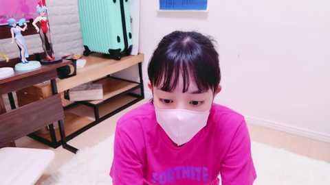 Media: A video of an Asian child with black hair, wearing a pink shirt and white face mask, sitting on a white rug in a brightly lit, minimalist room with light-colored walls.