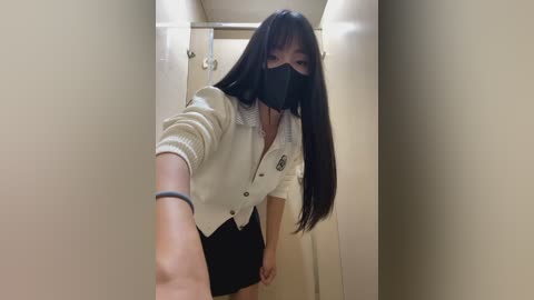 Media: Video of an Asian woman with long black hair, wearing a white button-up shirt, black mask, and black skirt, taking a selfie in a narrow, beige-tiled hallway with a door and a towel rack in the background.