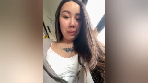 Media: Video of a young Asian woman with long, straight brown hair, wearing a white tank top with a grey cardigan, showing a detailed floral tattoo on her chest.