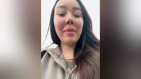 Media: Video of a young Asian woman with long, straight black hair, wearing a light gray hoodie. She has a tattoo on her chest, and her eyes are closed, giving a serene expression.