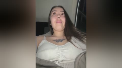 Media: Video of a young woman with long brown hair, wearing a white ribbed tank top, sitting in a dimly lit room. She has a tattoo of a black floral design on her chest.