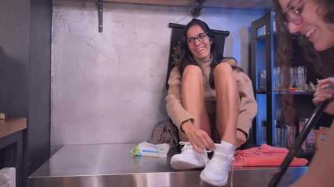 Media: Video of a woman in glasses, seated on a table, tying a white sneaker, with another woman in the background. Industrial setting with shelves and a blue light.