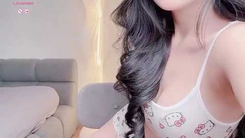 Media: Video of a young woman with long, wavy black hair, wearing a white tank top with Hello Kitty designs, sitting on a gray upholstered chair in a minimalist, modern room.