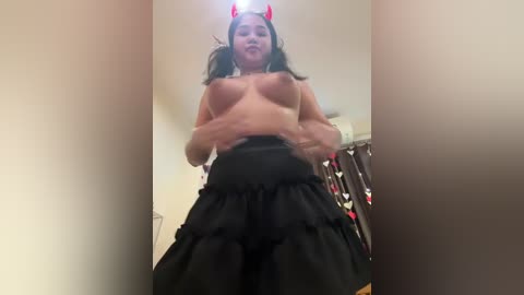 Media: A video shows a topless woman with medium breasts and long dark hair, wearing a red devil horn headband and a black ruffled skirt. She stands in a dimly lit room with a white wall and a hanging black robe.