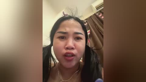 Media: Video of an Asian woman with long black hair in pigtails, wearing a black top, looking slightly surprised, in a dimly lit room with a brown curtain and air conditioning unit in the background.