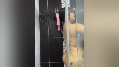 Media: Video of a naked woman with long black hair, wearing a pink hairband, standing in a black-tiled shower, viewed through a frosted glass door, revealing her bare back and buttocks.