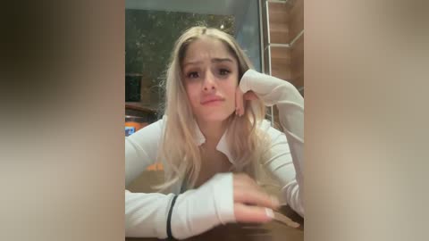 Media: Video of a young, fair-skinned, blonde woman with long hair, wearing a white cardigan, resting her head on her hand in a thoughtful manner. Background shows a wooden table and a staircase.