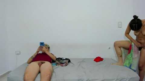 Media: Video of two women in a bedroom: one topless with fair skin and blonde hair, taking a selfie; another, also topless with fair skin and dark hair, bending over to pick up clothes.