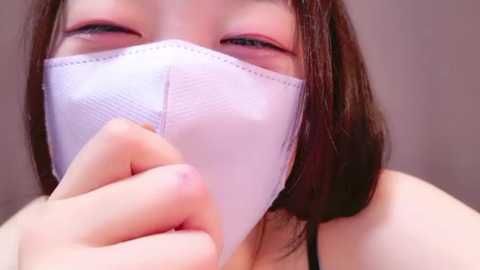 Media: Video of a young woman with fair skin, wearing a white surgical mask, holding it to her mouth, brown hair partially visible. Background is blurred.