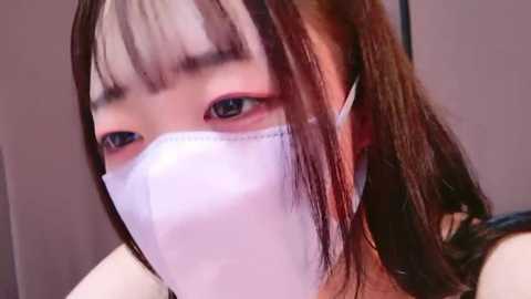 Media: A close-up video of a young woman with light skin and long, straight brown hair, wearing a white surgical mask and a black top. She has bangs and looks slightly to the left.