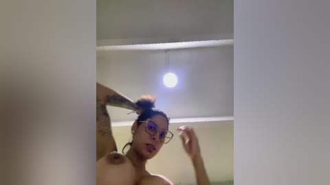 Media: Video of a topless, light-skinned woman with medium-length, dark hair tied in a bun, wearing glasses, and tattoos on her arms, standing indoors with a white ceiling and light fixture.